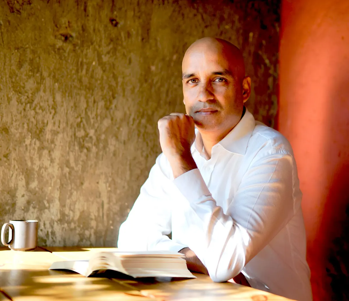 Greg Chapman sitting at a wooden table with an open book and a coffee mug, chin resting on his hand