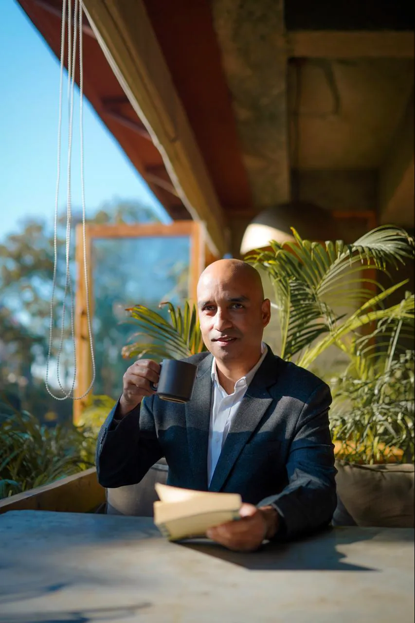 Greg Chapman at a terrace table with a coffee mug and a book, surrounded by plants