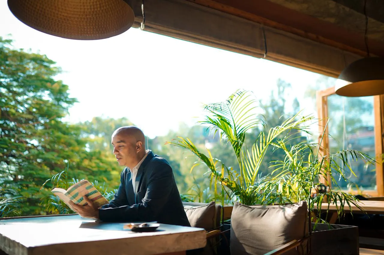 Greg Chapman reading a book at a terrace table, trees and plants behind him