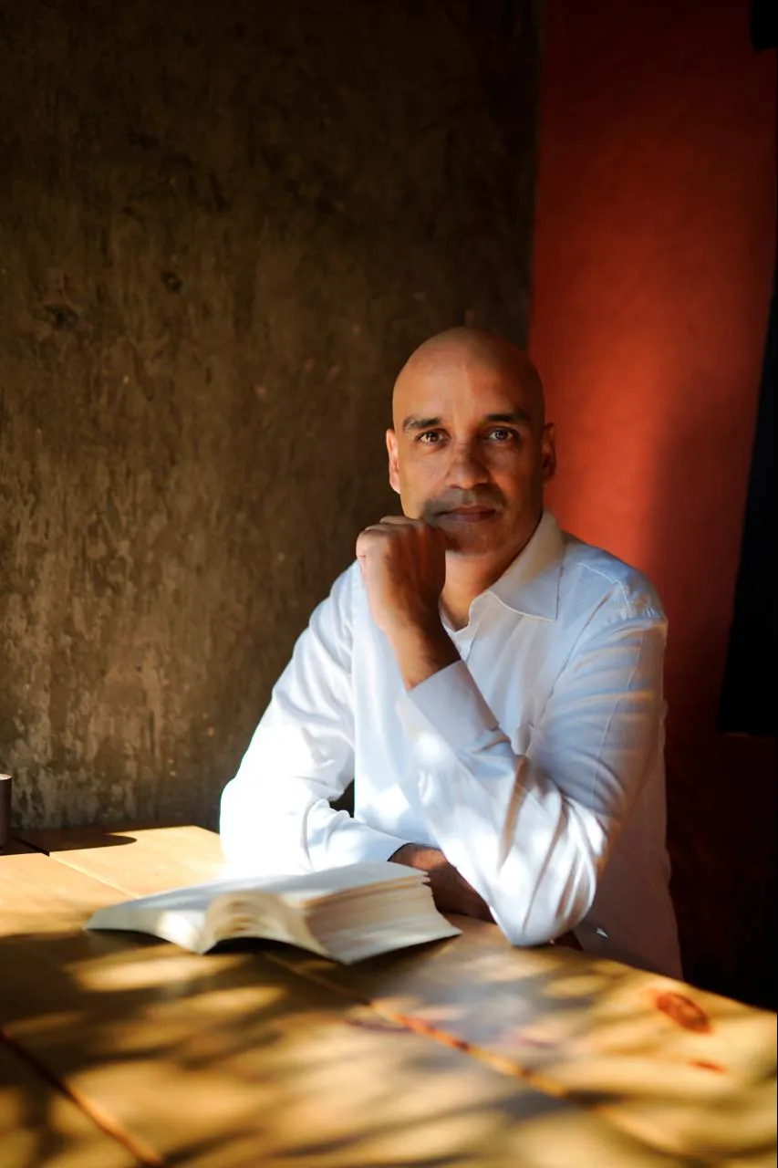 Greg Chapman in a white shirt at a wooden table beside a red wall, chin resting on his hand, an open book in front of him